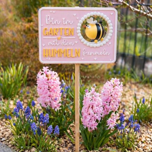 Rosafarbenes Gartenschild mit Hummelmotiv und Spruch zwischen blühenden Frühlingsblumen